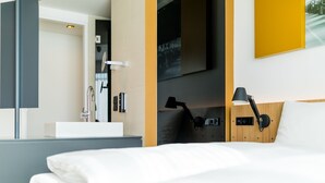 Une chambre d’hôtel moderne avec un grand lit, une table de chevet avec une lampe, un miroir et une vue sur le paysage urbain.