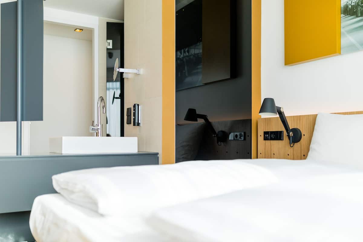 Un hotel moderno con una cama grande, una mesita de noche con lámpara, un espejo y vistas a la ciudad.