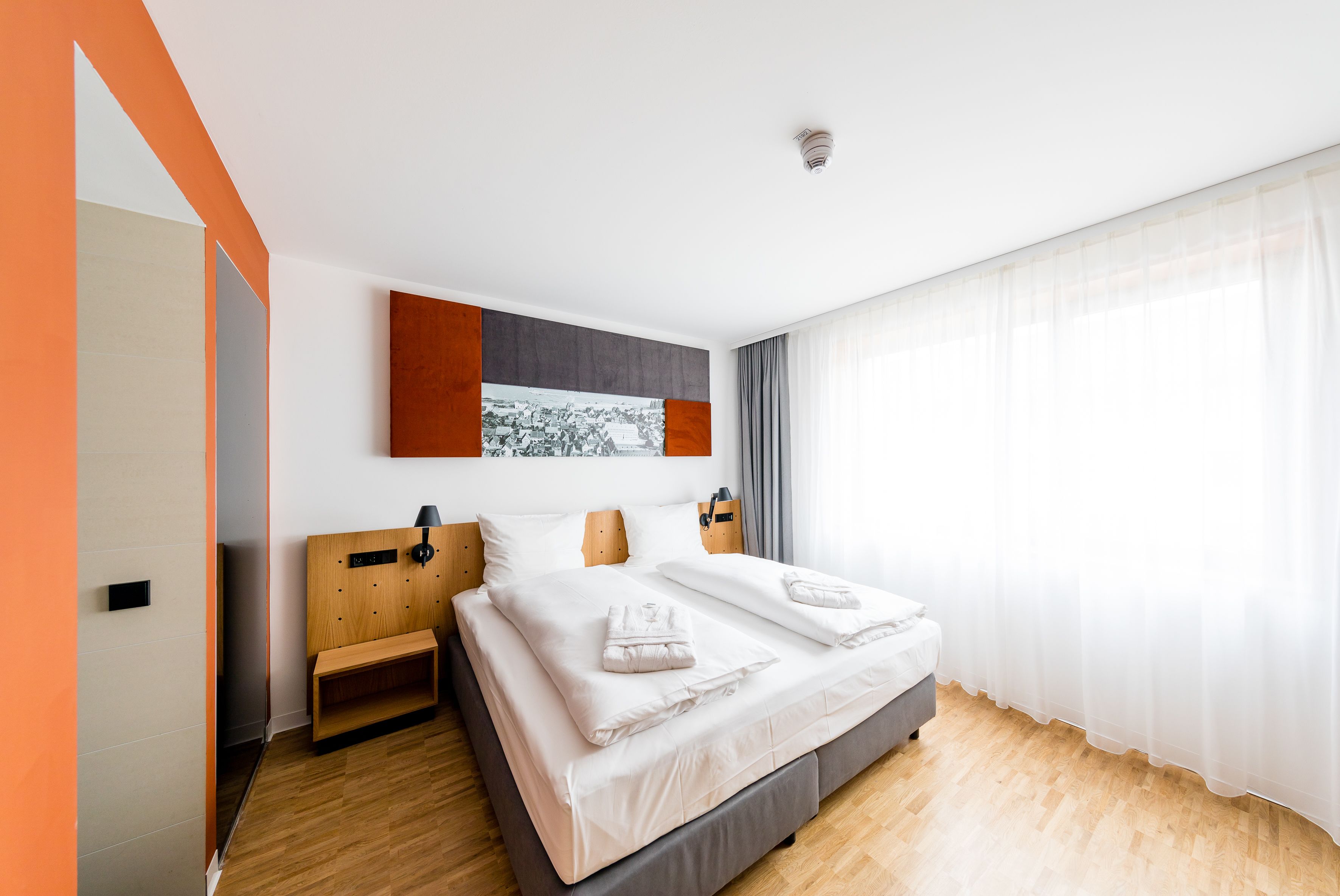 A modern hotel room with a large bed, a wooden headboard, a bench, and a large window with sheer curtains.