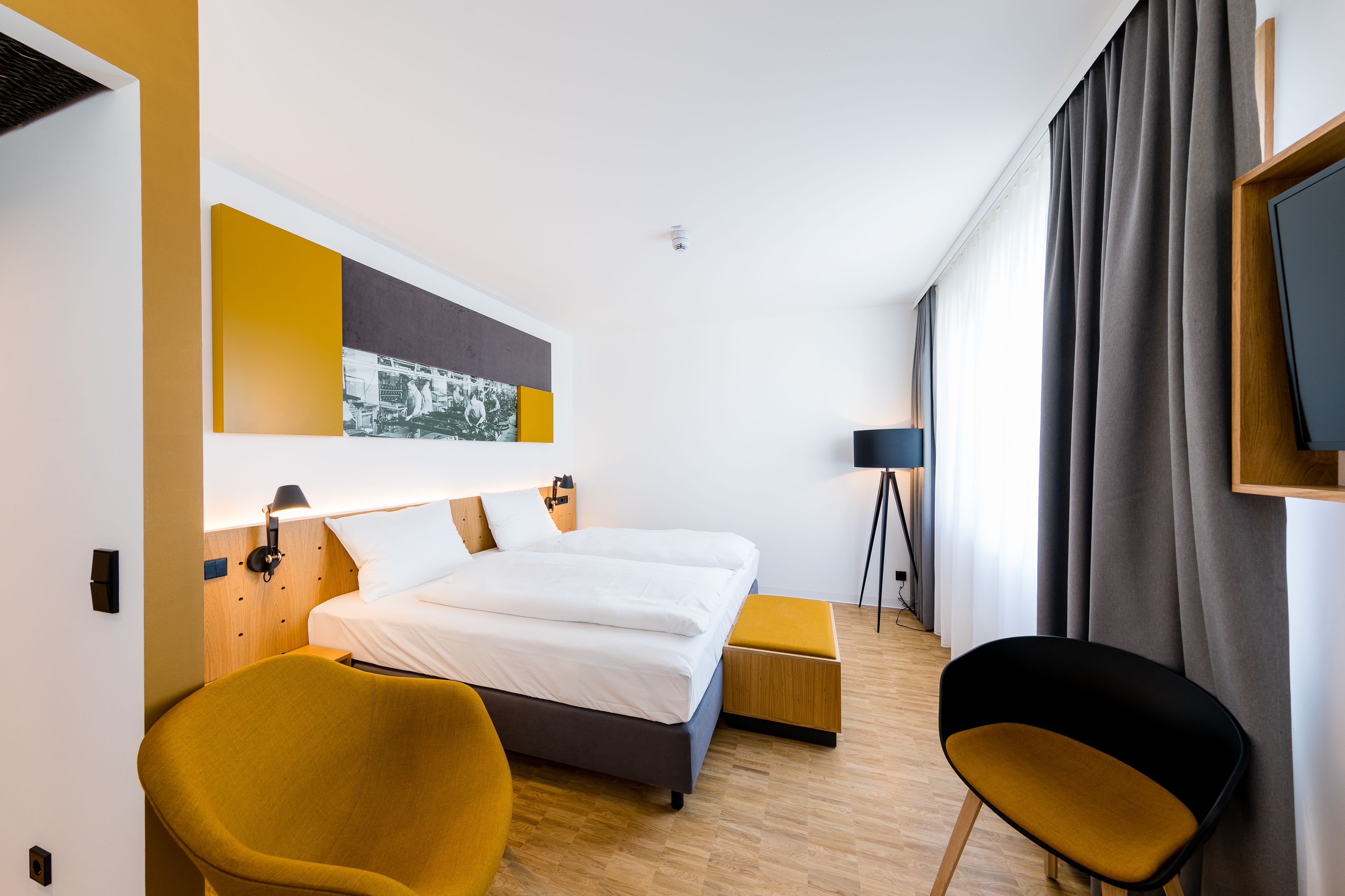 Una habitación de hotel moderna con una cama grande, un sillón amarillo, una silla negra y un cuadro en la pared.