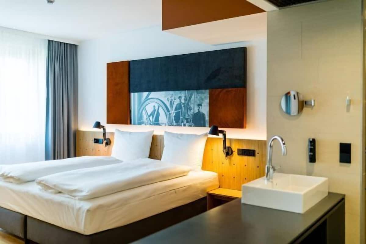 Habitación de hotel con cama doble, mesita de noche, lavamanos moderno y dispensador de toallas en la pared.