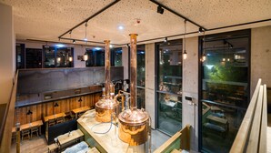 Un espace bar moderne, doté d’un équipement de distillation en cuivre, de tables en bois et d’un plafond haut orné d’un motif moucheté.