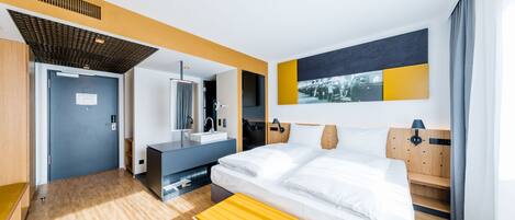 A modern hotel room with a large bed, a desk with a computer, a bathroom, and a view of the outdoors.