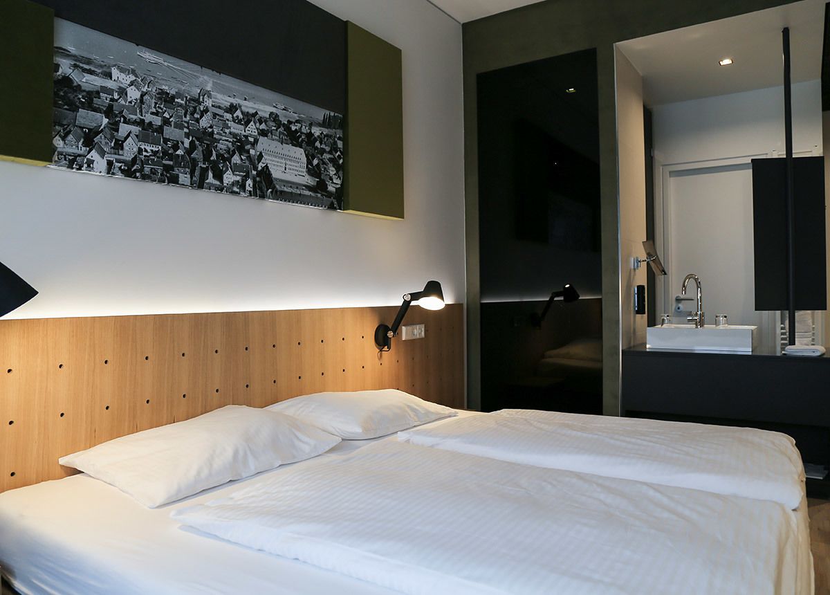 A modern hotel room with a wooden headboard, a large bed, a nightstand with a lamp, a bathroom with a mirror and sink, and a poster on the wall.