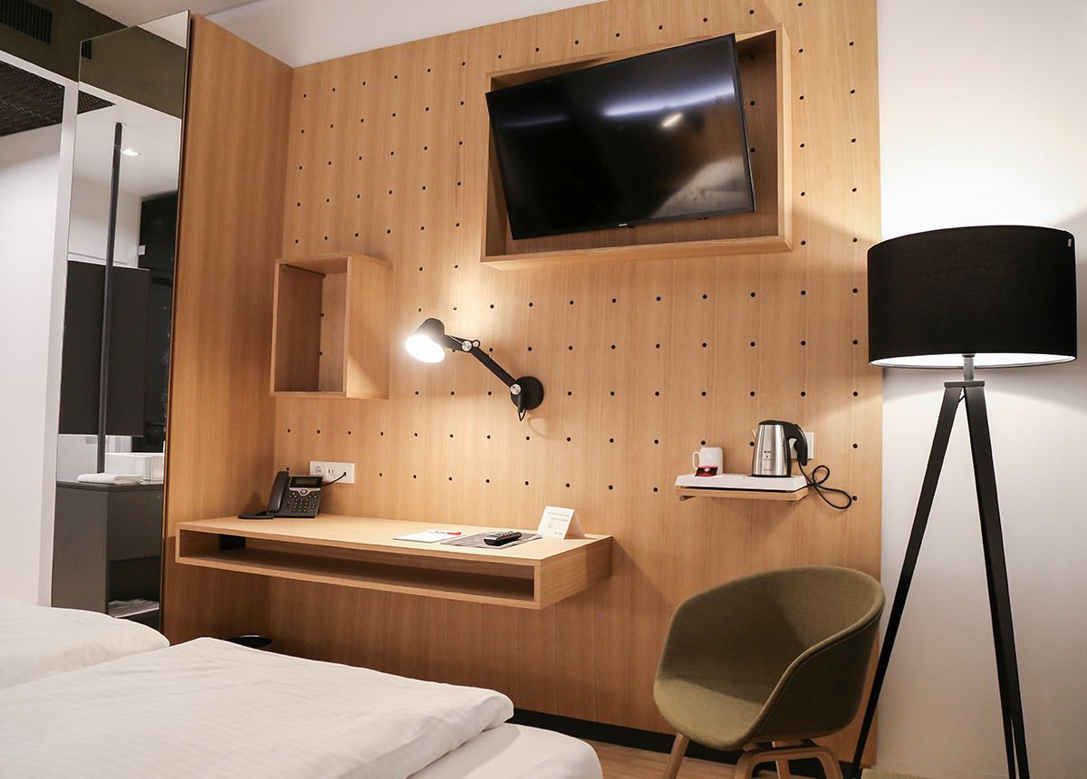 A hotel room with a wooden wall, a flat-screen TV, a desk with a lamp, a chair, and a bedside table.