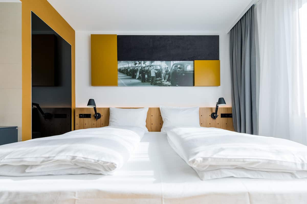 Habitación de hotel con una cama grande, dos mesitas de noche, una obra de arte colgada en la pared y un ventanal con cortinas.