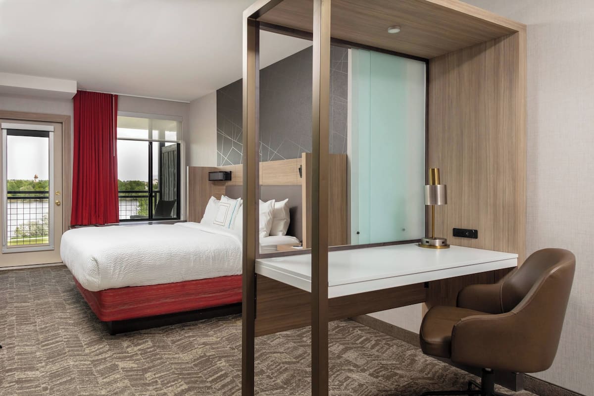 A hotel room with a bed, a desk, a chair, a window with curtains, and a view of greenery outside.