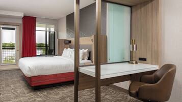 A hotel room with a bed, a desk, a chair, a window with curtains, and a view of greenery outside.