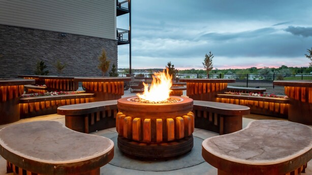 An outdoor seating area with a central fire feature surrounded by wooden benches and tables. - SpringHill Suites by Marriott Great Falls (Great Falls)