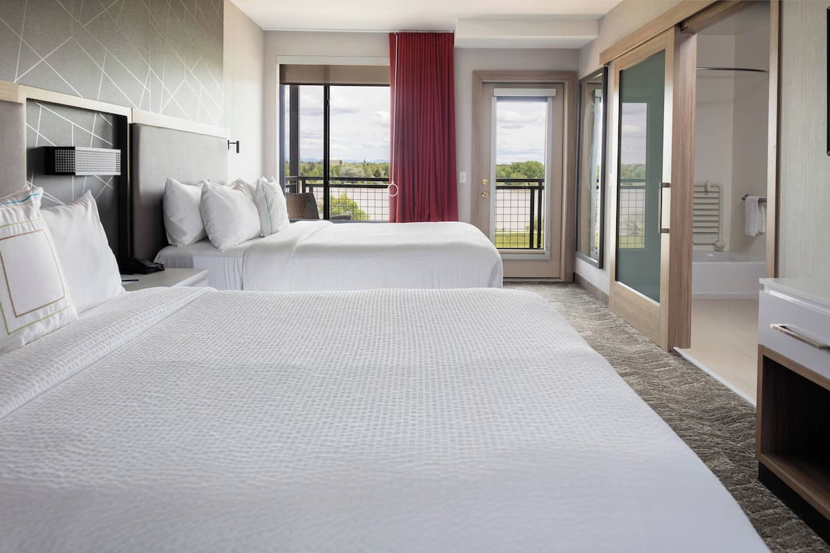 A hotel room with two beds, a large window with a view, a sliding door to a balcony, and a bathroom with a bathtub.
