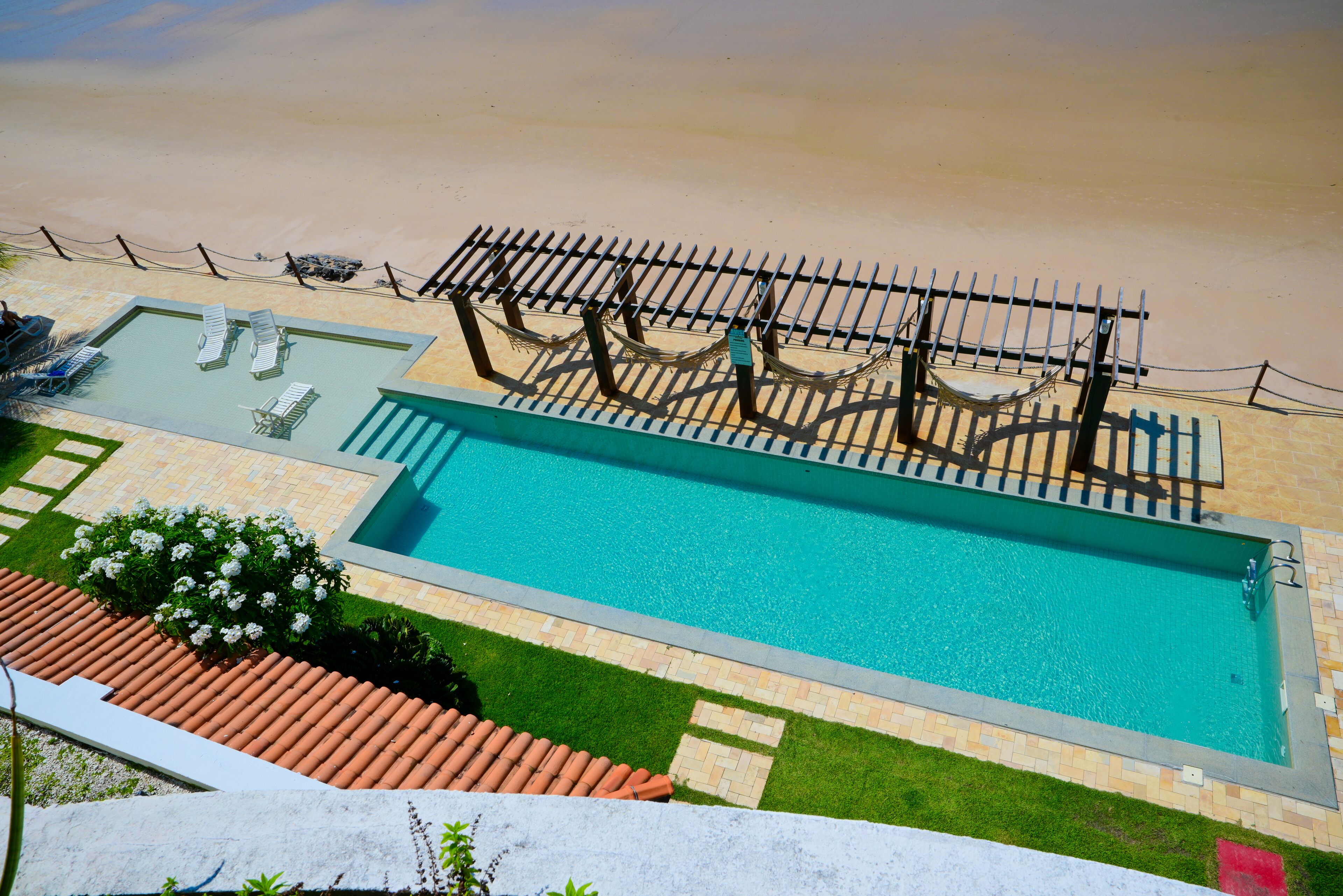 A swimming pool with a sun deck, surrounded by a sandy beach and a fenced area with lounge chairs.