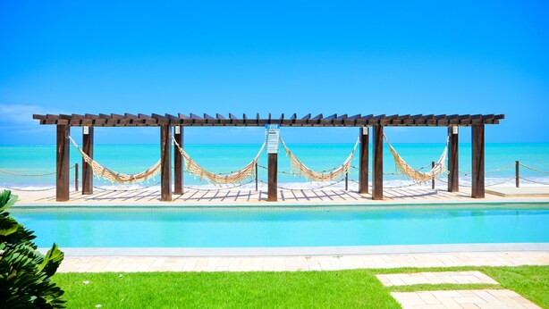 A wooden pergola with a hammock over a pool, surrounded by a clear blue sky and ocean view. - Waterfront Apart Hotel (Maceió)