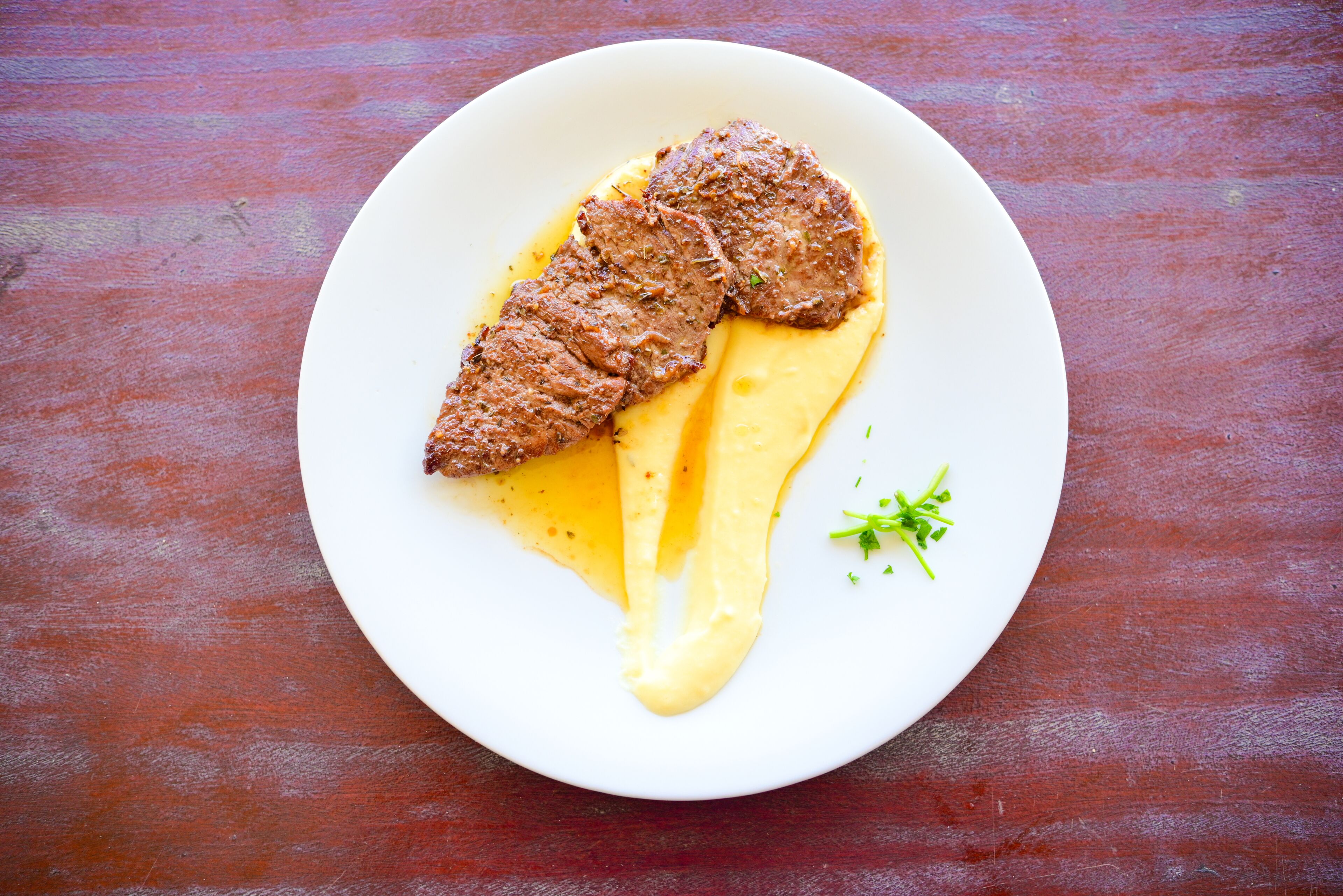 Une assiette blanche contenant un morceau de viande et de la purée de pommes de terre, posée sur une table en bois.