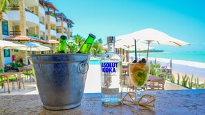 A table with a bucket containing bottles, a bottle of Absolut Vodka, and a cocktail glass with a straw, overlooking a beach with umbrellas and a building in the background. - Waterfront Apart Hotel (Maceió)