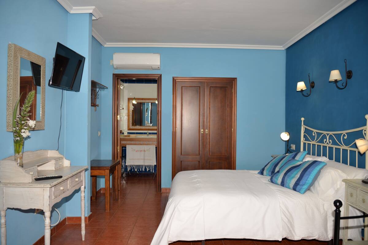 Ein Schlafzimmer mit Bett, Schreibtisch mit Stuhl, Fernseher, Kleiderschrank und einem durchgegangenen Tür erkennbaren Badezimmer.