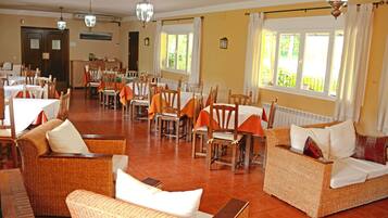Un comedor amplio con varias mesas y sillas, mobiliario de mimbre y grandes ventanales que dejan entrar luz natural.