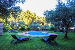 Un patio trasero con piscina circular, rodeado de césped y árboles. Hay sillas de descanso y un adorno circular rojo.