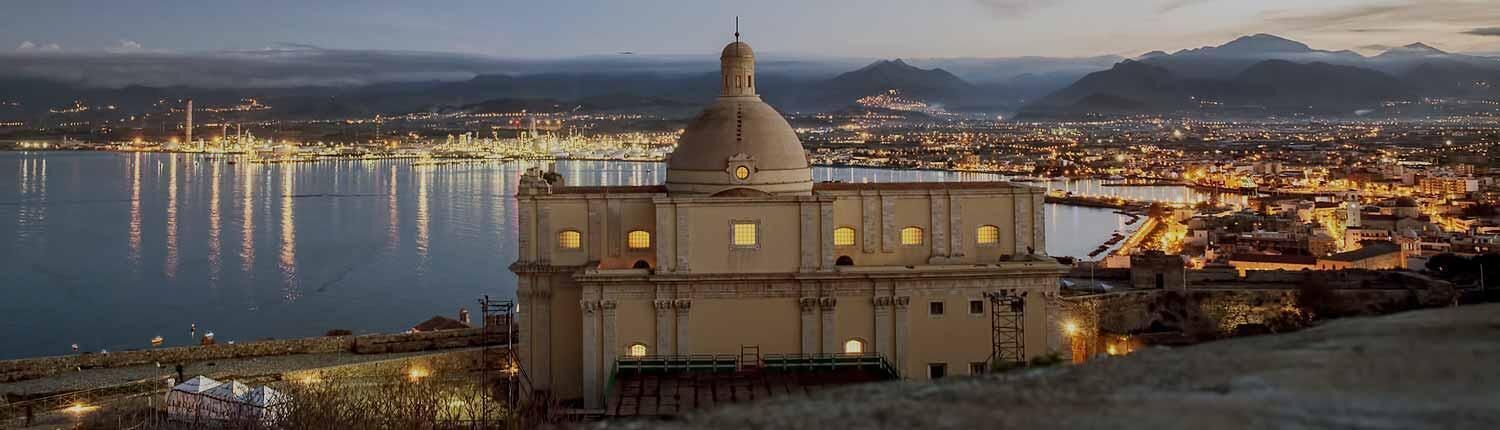 A historic building with a dome, situated by a body of water with city lights in the background.
