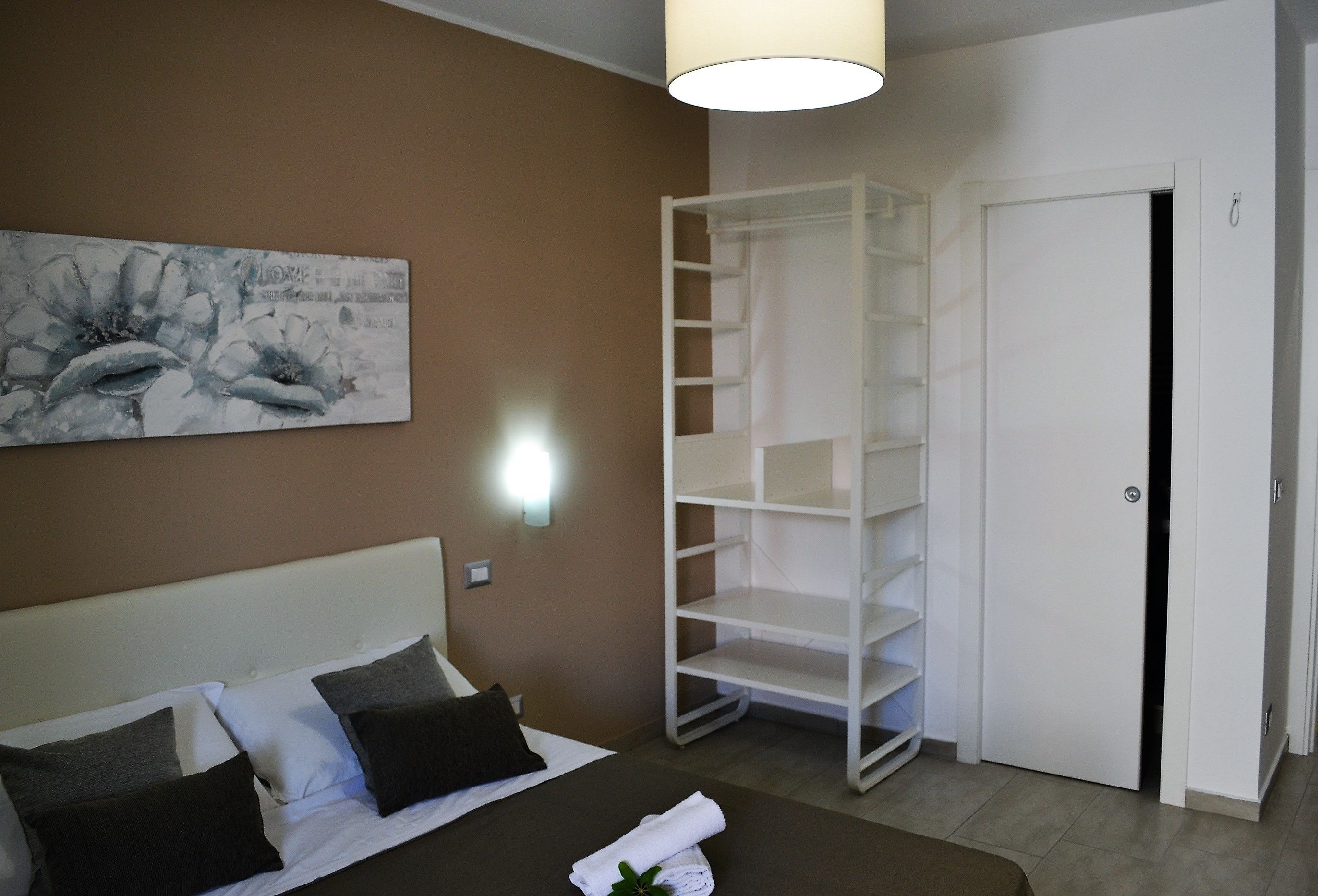 A hotel room with a bed, a white wardrobe, a white shelving unit, and a wall-mounted light.