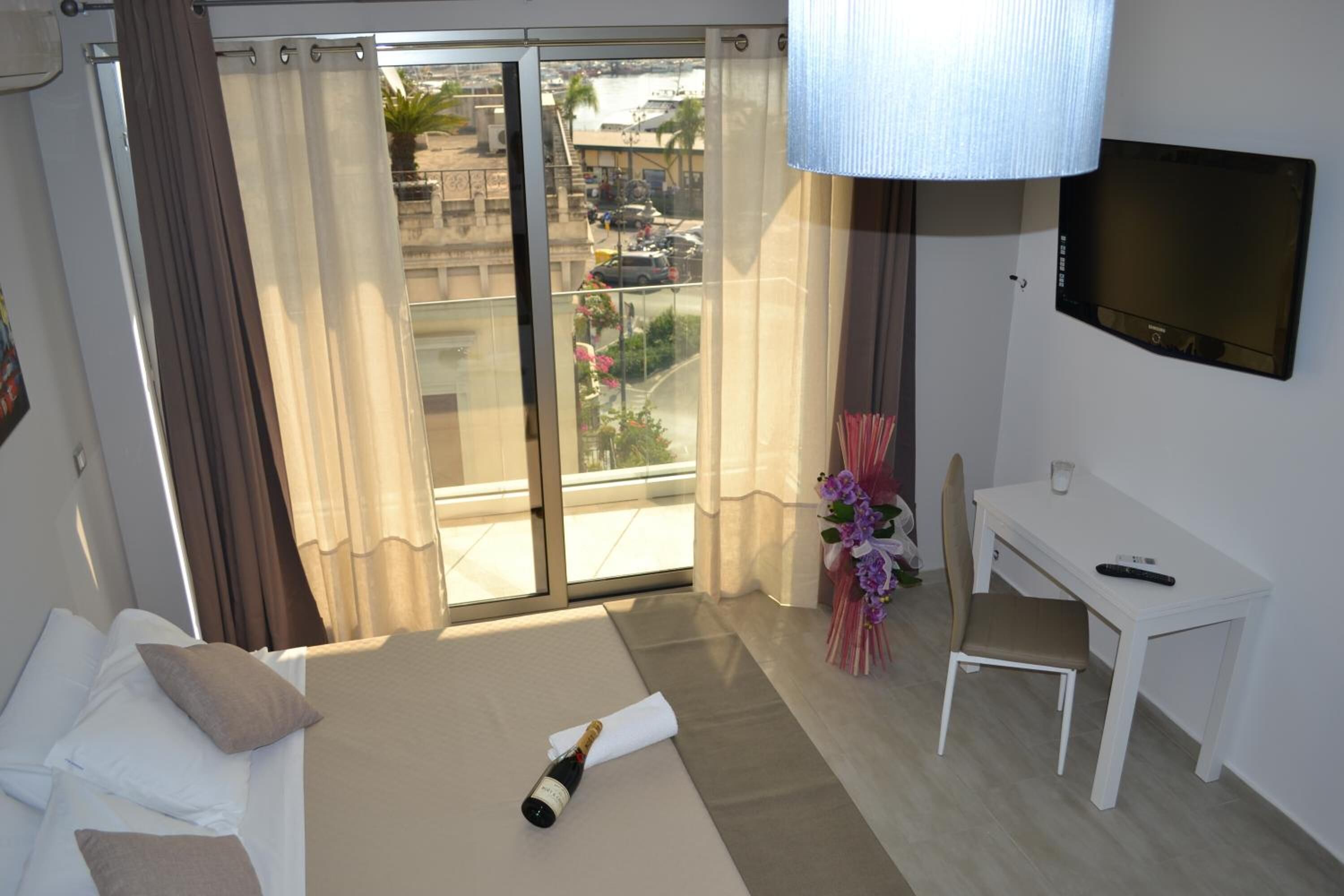 A hotel room with a bed, a desk, a chair, a TV, and a balcony with a view of the city.
