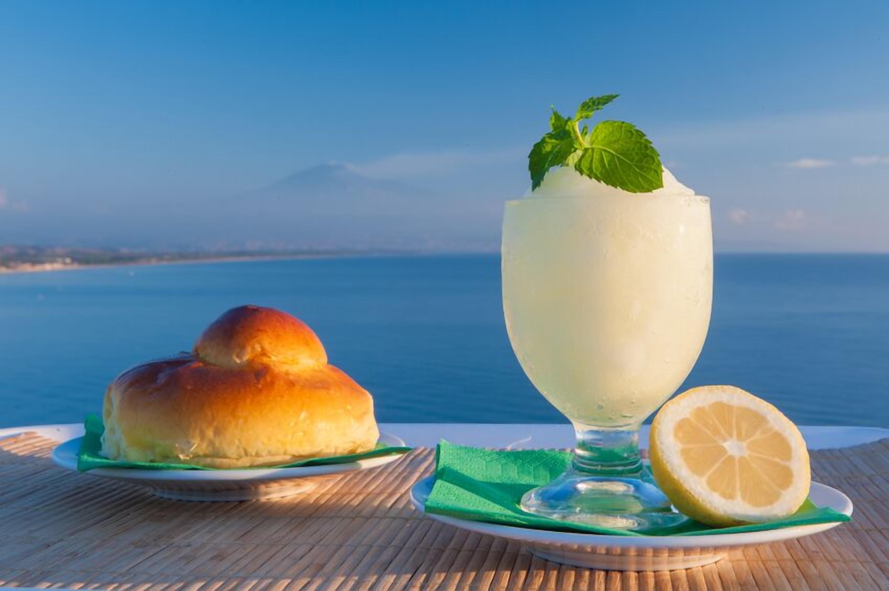 A table with a glass of a white beverage garnished with a mint leaf, a half lemon, and a roll of bread on a plate.