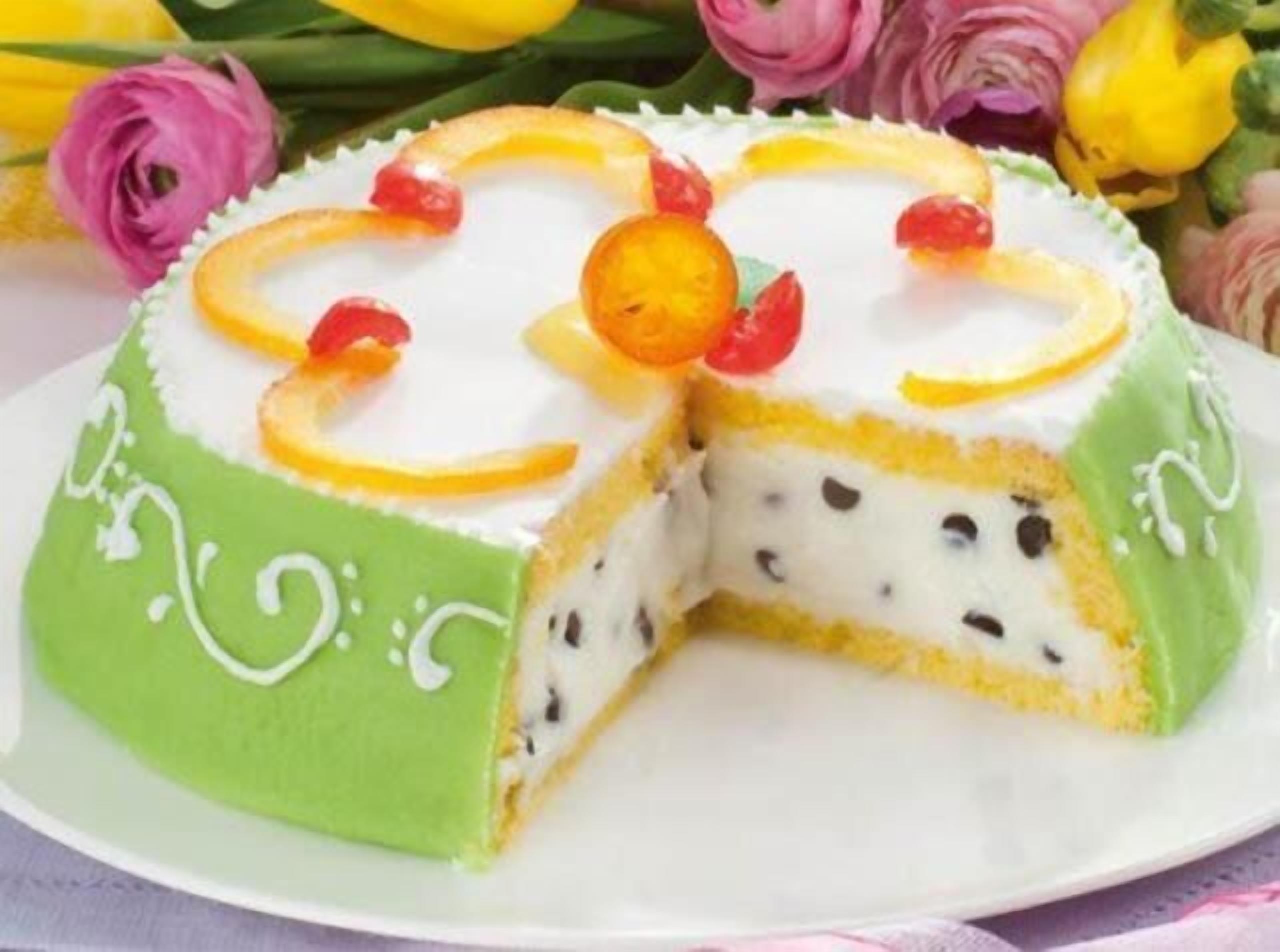 Een plak gebak met een groene buitenkant en een witte binnenkant, versierd met fruitreepjes en chocoladechips.