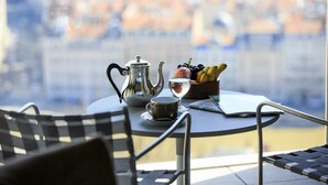 Una mesa metálica redonda con una cafetera, una taza, un vaso de agua, un cuenco de fruta y un menú en un balcón con vistas a un paisaje urbano.