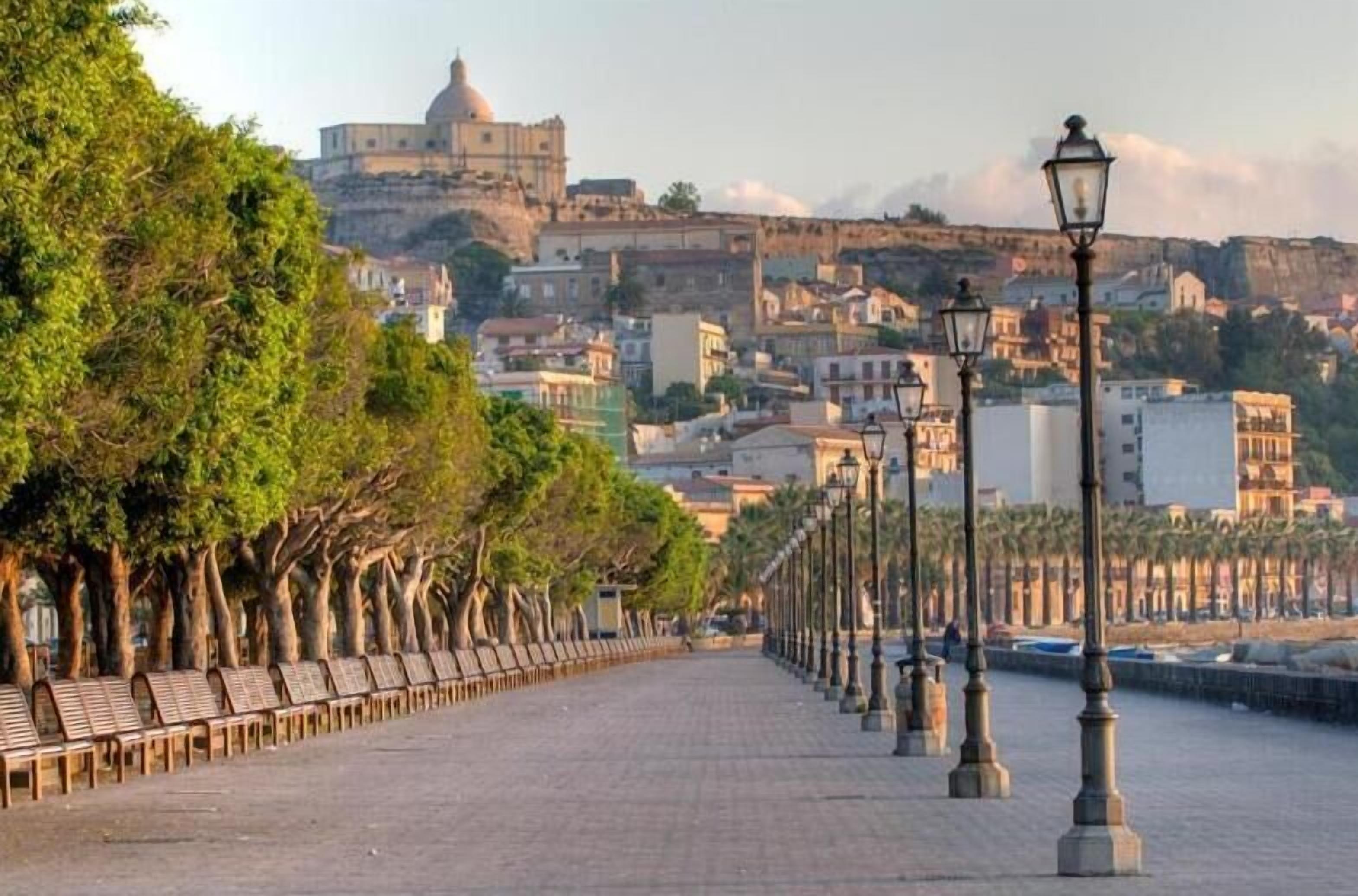 Un chemin piétonnier, bordé de bancs et de lampadaires, mène à un bâtiment historique surmonté d’un dôme.