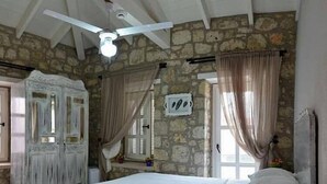 A bedroom with a stone wall, a bed with white linens, a ceiling fan, and a window with curtains. - Simira Hotel (Cesme)