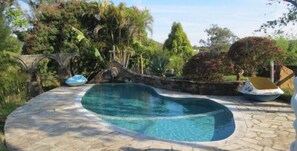 Una piscina con borde de piedra, rodeada de exuberante vegetación y un puente de piedra.