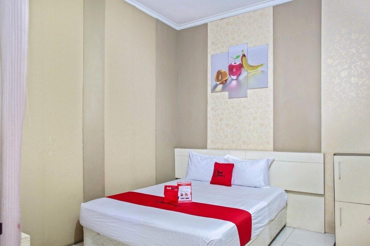 A hotel room with a bed, a red and white bedspread, a red pillow, and a wall with a fruit painting.