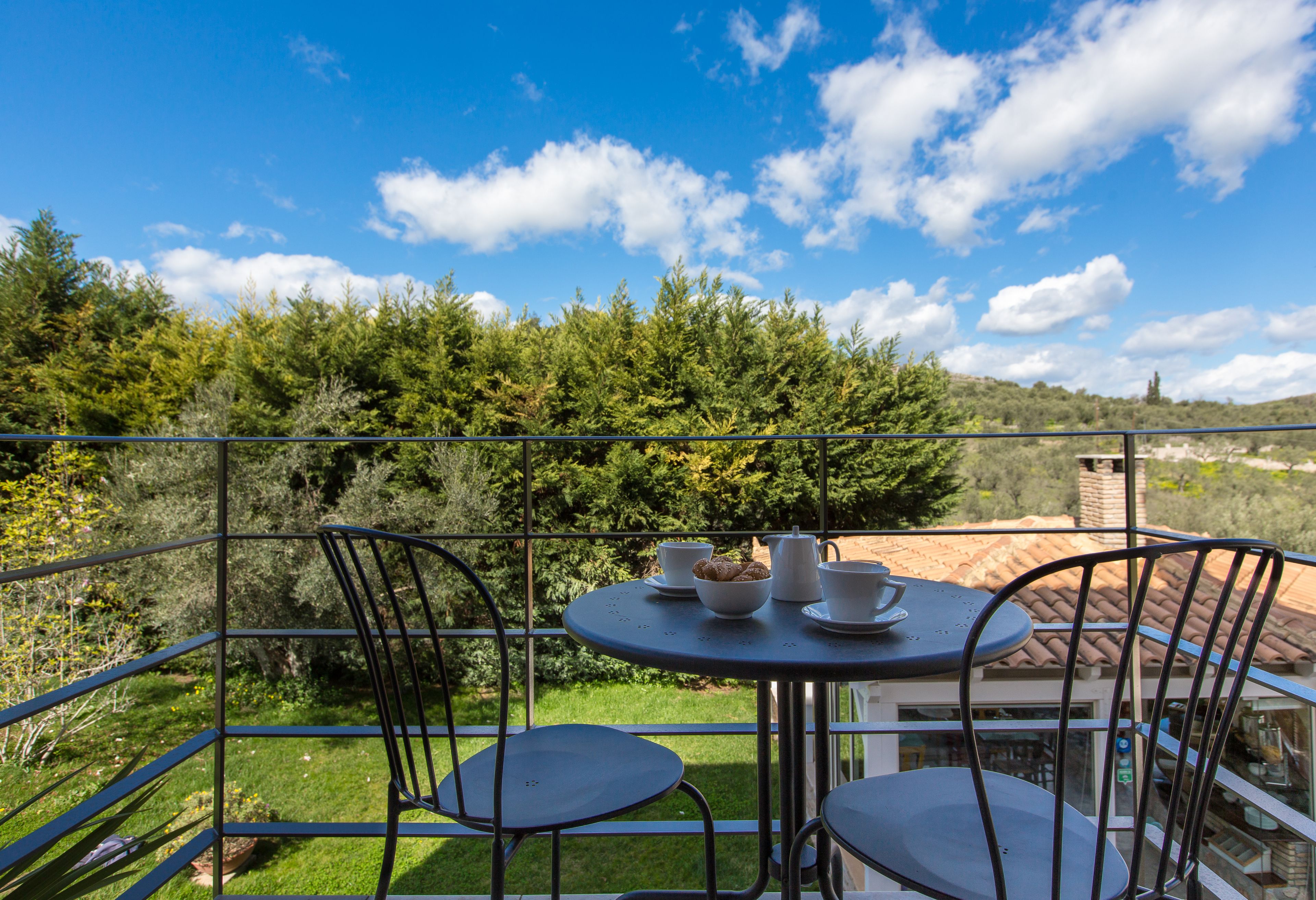 A balcony with a table set for two, surrounded by chairs, overlooking a lush green landscape and a clear blue sky.