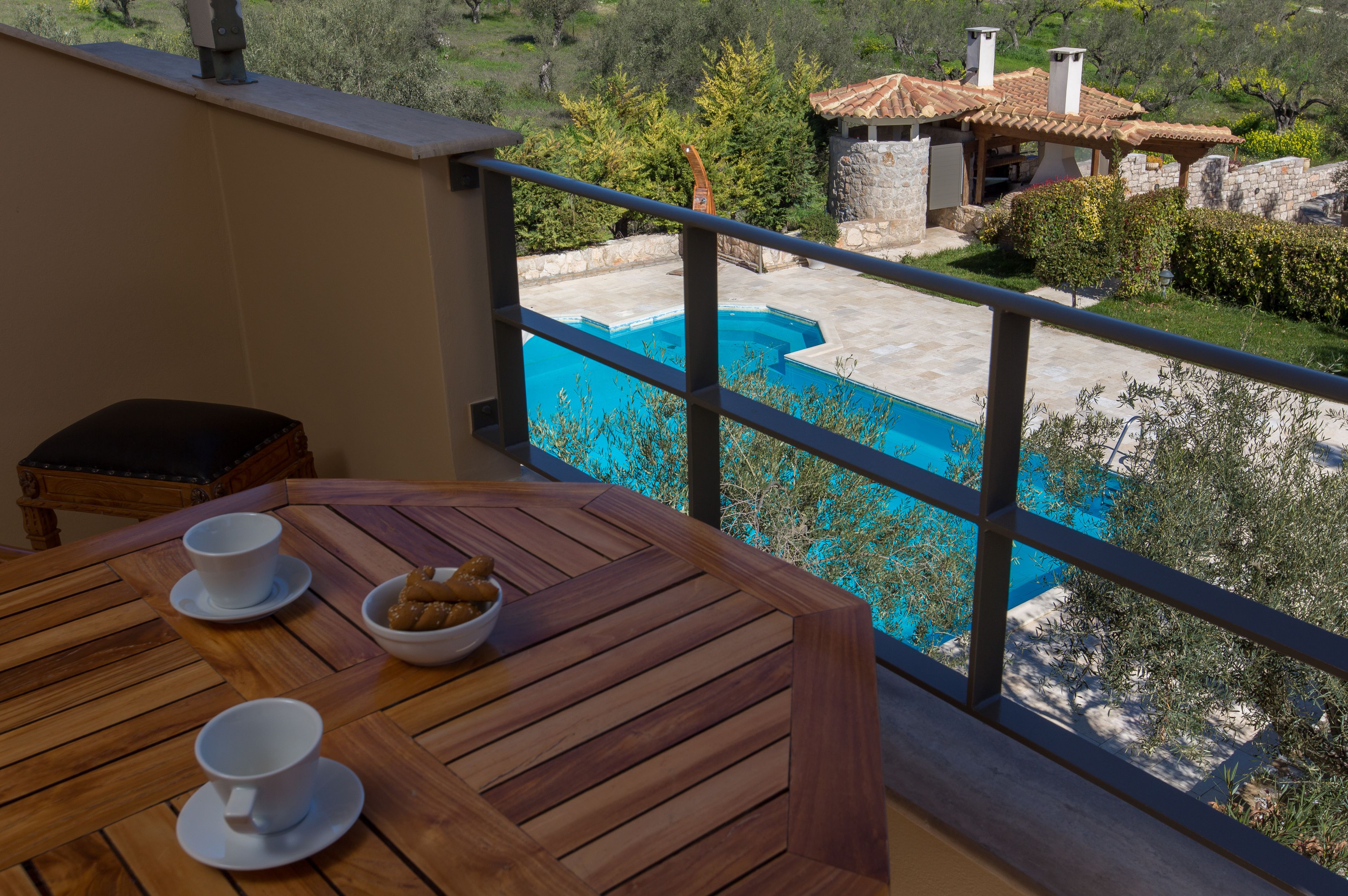 A wooden table with two white cups and a bowl of snacks, overlooking a swimming pool and a stone building with a tiled roof.