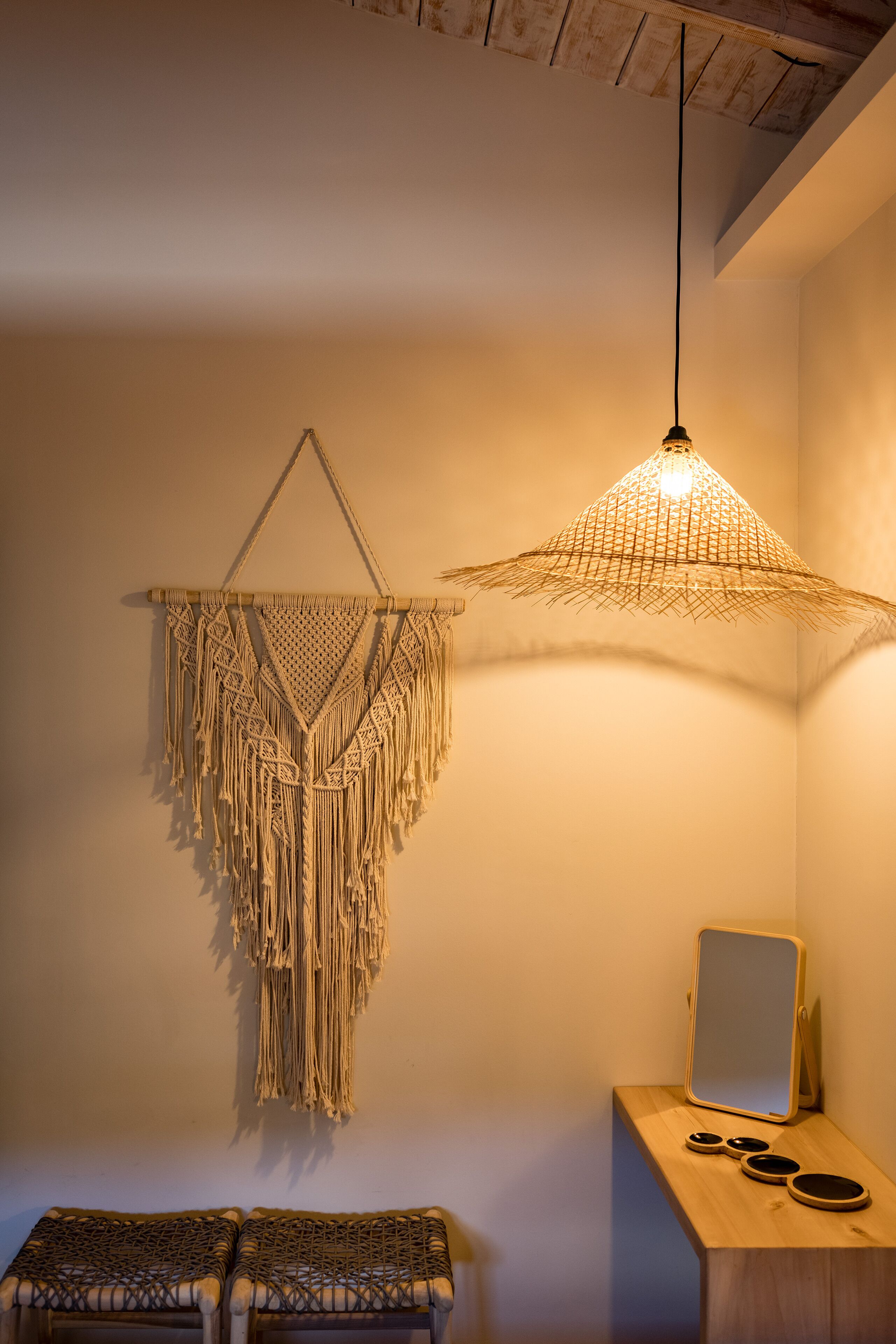 Ein Zimmer mit Wandbehang aus Makramee, einer Pendelleuchte, einer Holzbank und einem kleinen Tisch mit Spiegel und Dekoration.