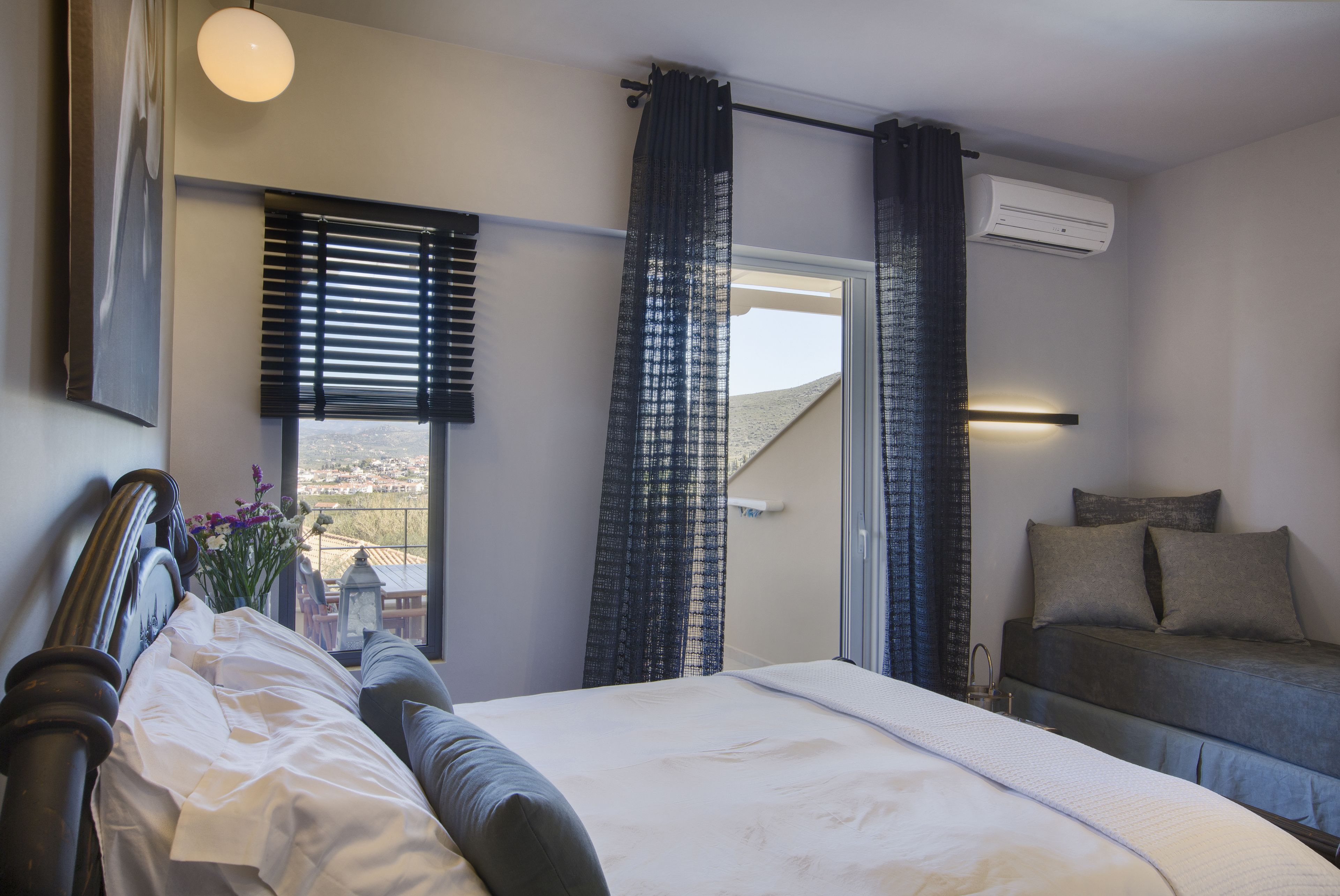 A bedroom with a bed, a sofa, a window with blinds, and an air conditioning unit.