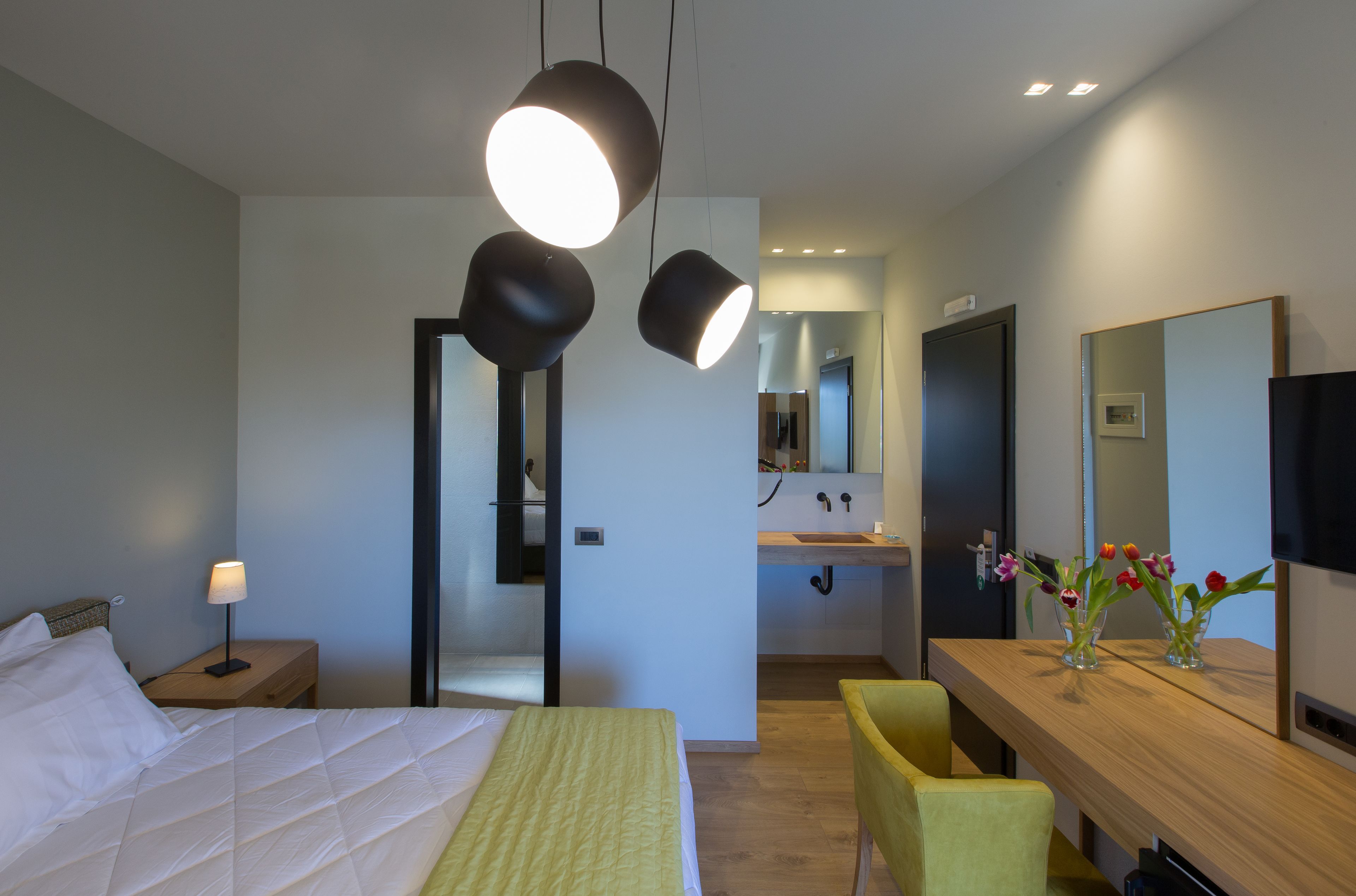 Une chambre d’hôtel moderne, équipée d’un lit, d’une table de chevet, d’une lampe et d’un bureau en bois agrémenté d’un miroir et d’un vase de fleurs.