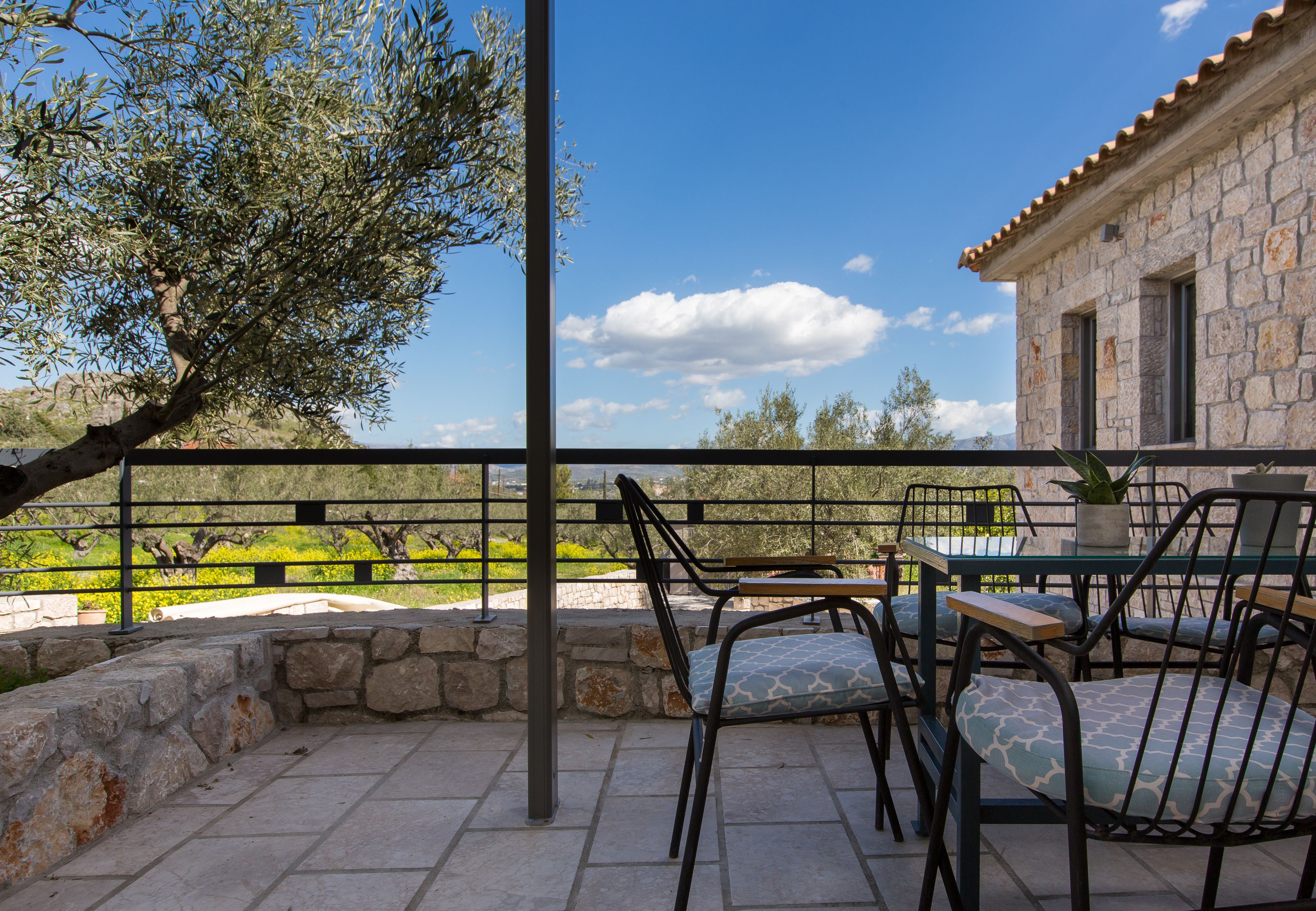 Un patio al aire libre con suelo de piedra, sillas de metal y una mesa. Se ve un edificio de piedra y un árbol al fondo.