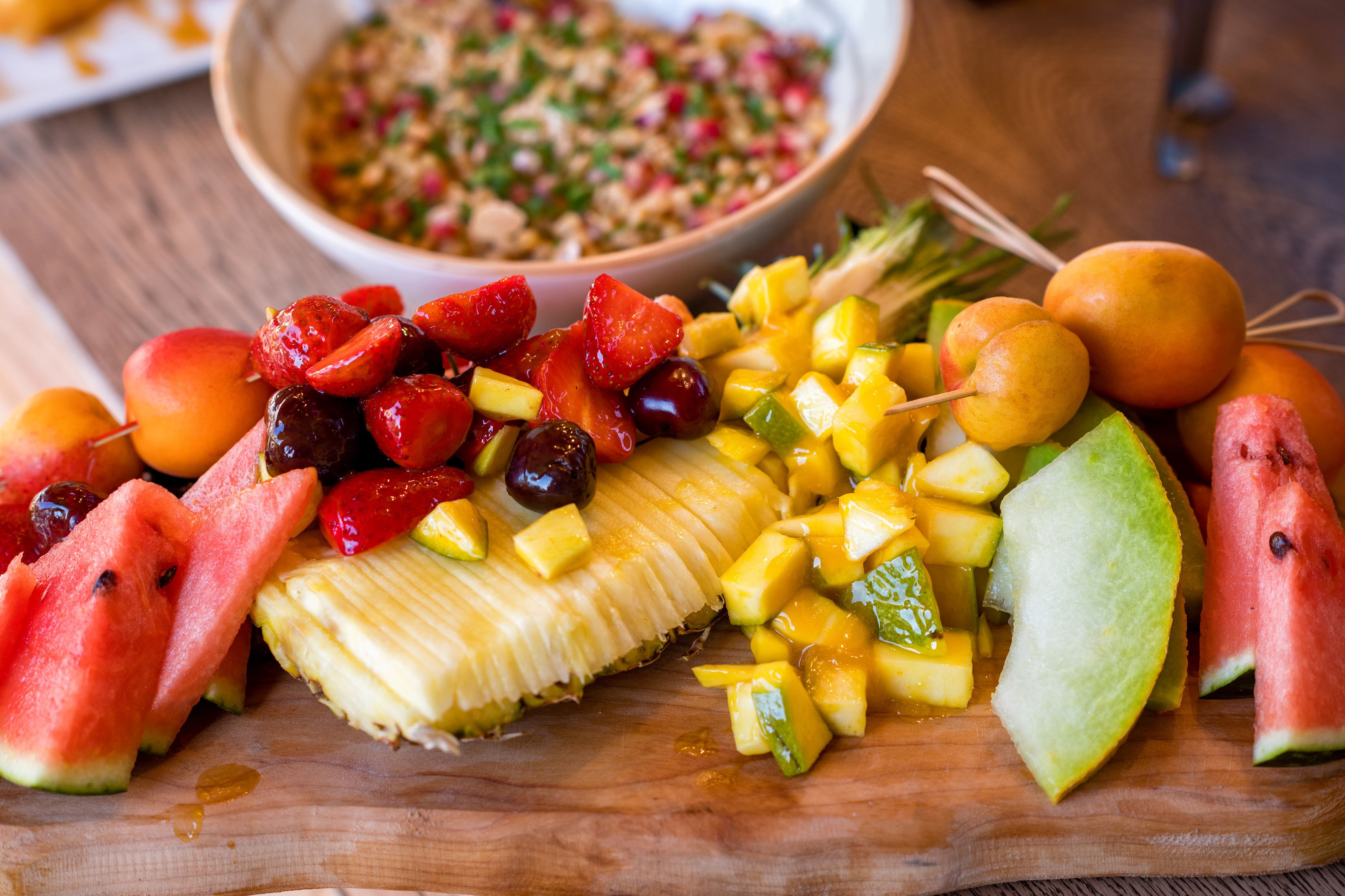 A wooden cutting board with sliced fruits and a bowl of salad in the background.