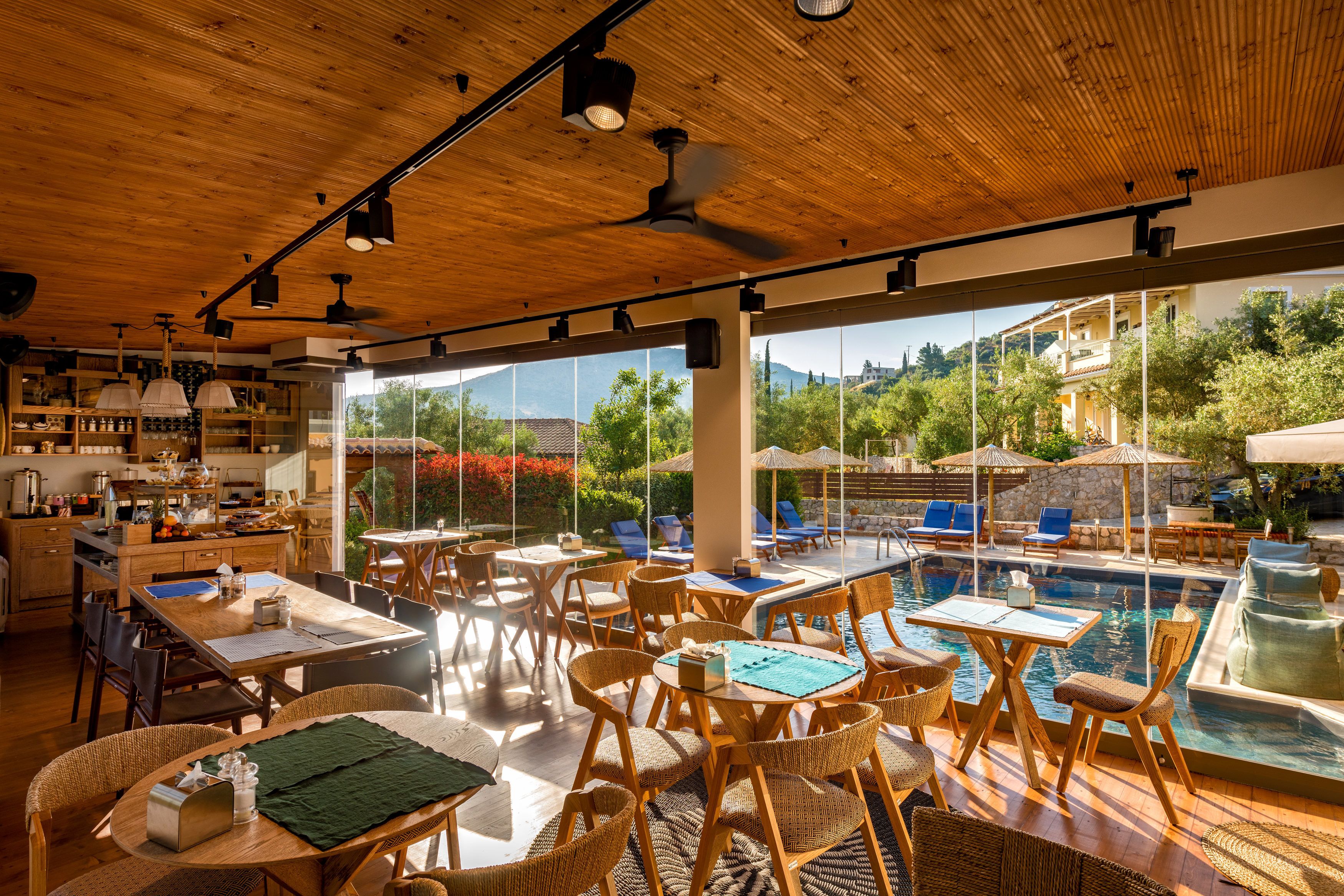 A spacious indoor area with wooden ceiling and large windows overlooking a pool and lush greenery.