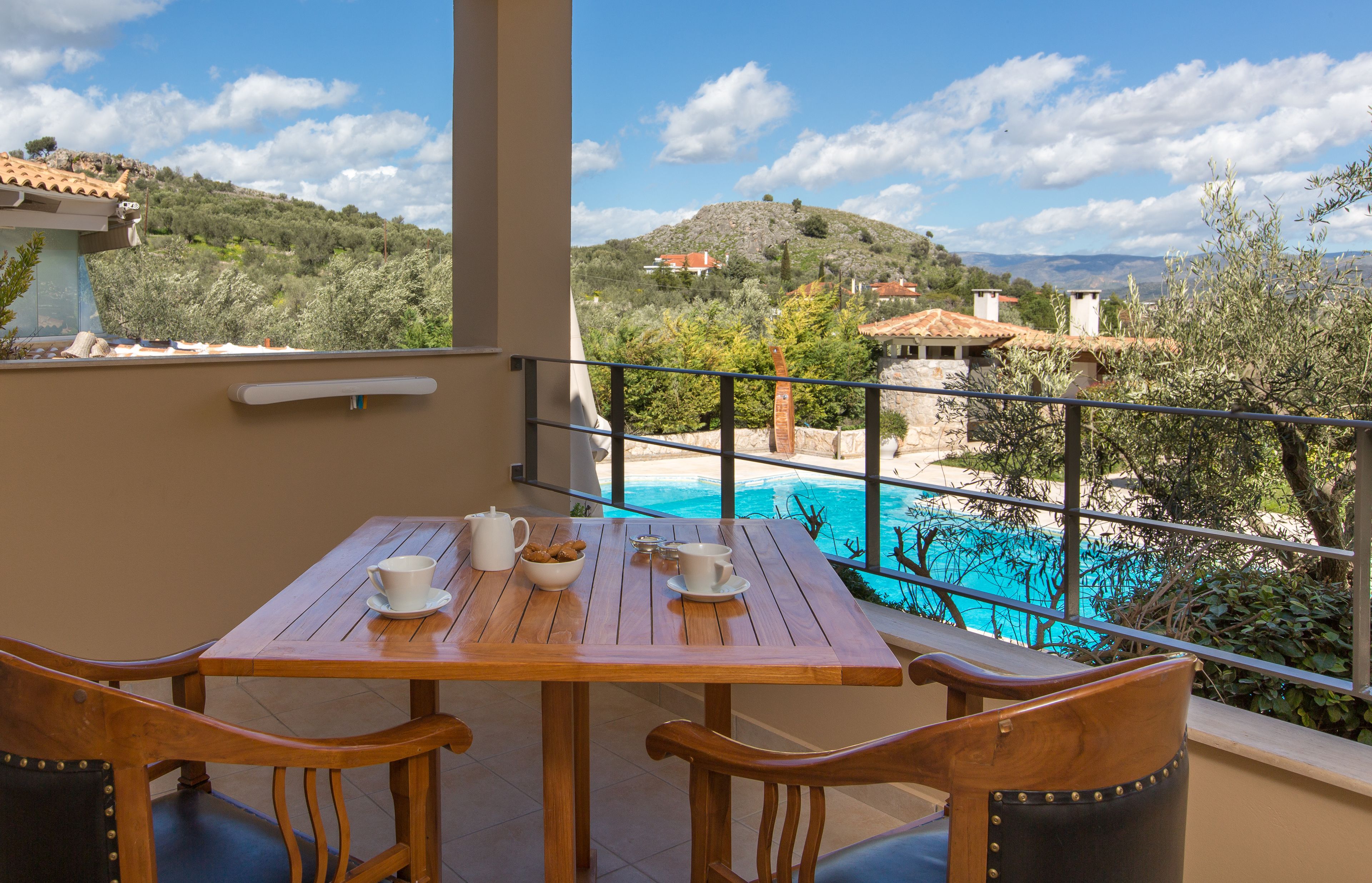 A wooden table with chairs and a view of a swimming pool and hillside houses.