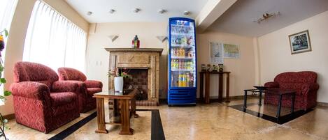 Un vestíbulo amplio con chimenea, sillones estampados de color rojo, un dispensador de bebidas azul y un piso de baldosas.