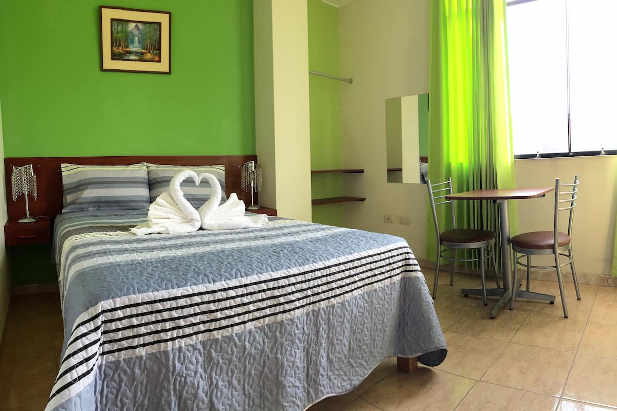 A hotel room with a bed, a painting on the wall, a green curtain, and a small table with two chairs.