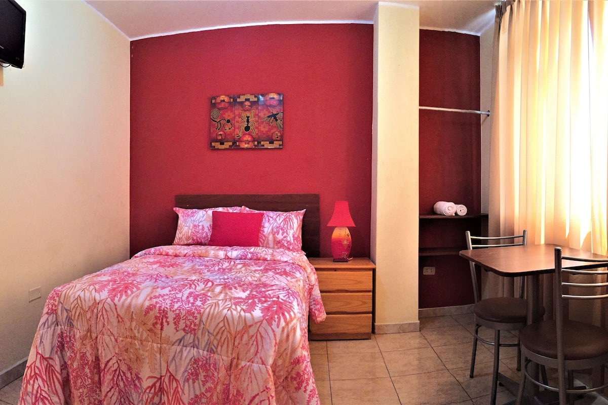A hotel room with a bed, a nightstand, a lamp, a chair, and a small table.