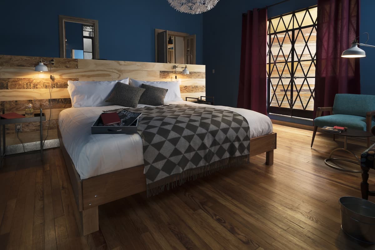 Une chambre moderne avec une tête de lit en bois, un grand lit, un plancher en bois, une fenêtre ornée d’un motif géométrique et une chaise bleue.