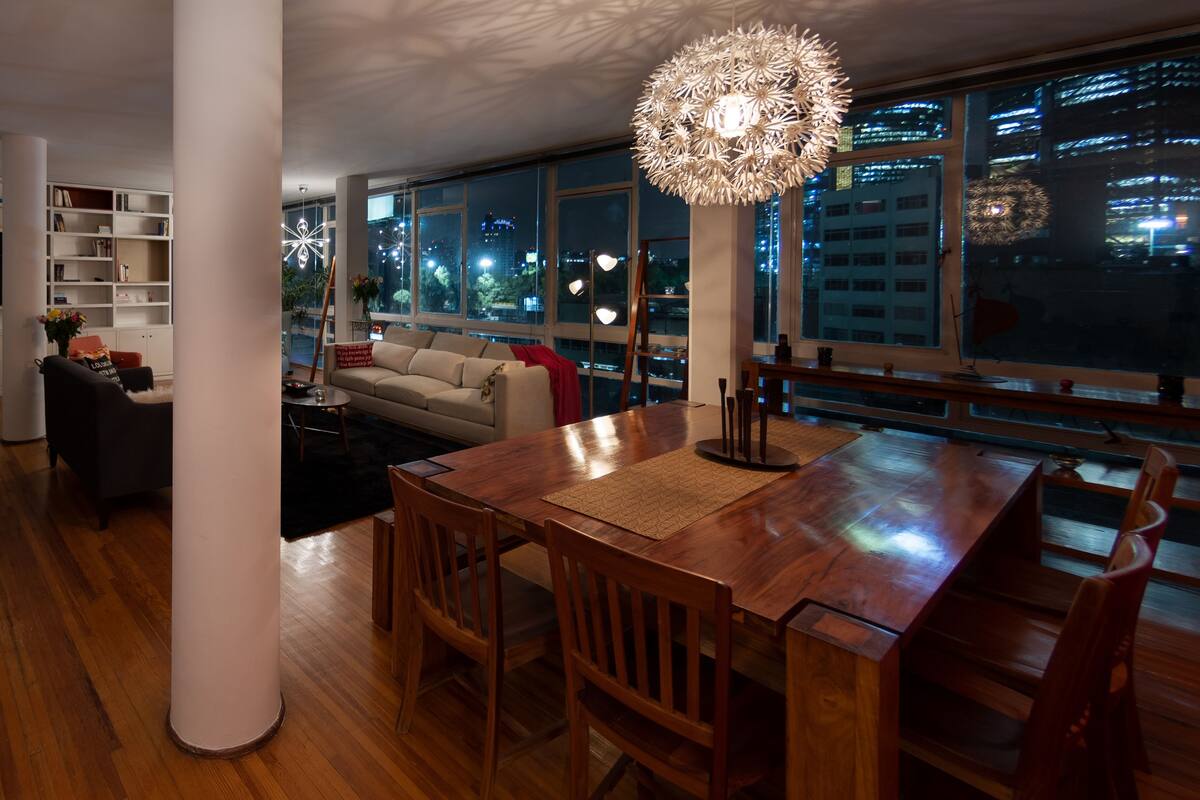 Un vaste espace de vie comprenant une table à manger en bois, un canapé et une grande fenêtre offrant une vue sur la ville la nuit.