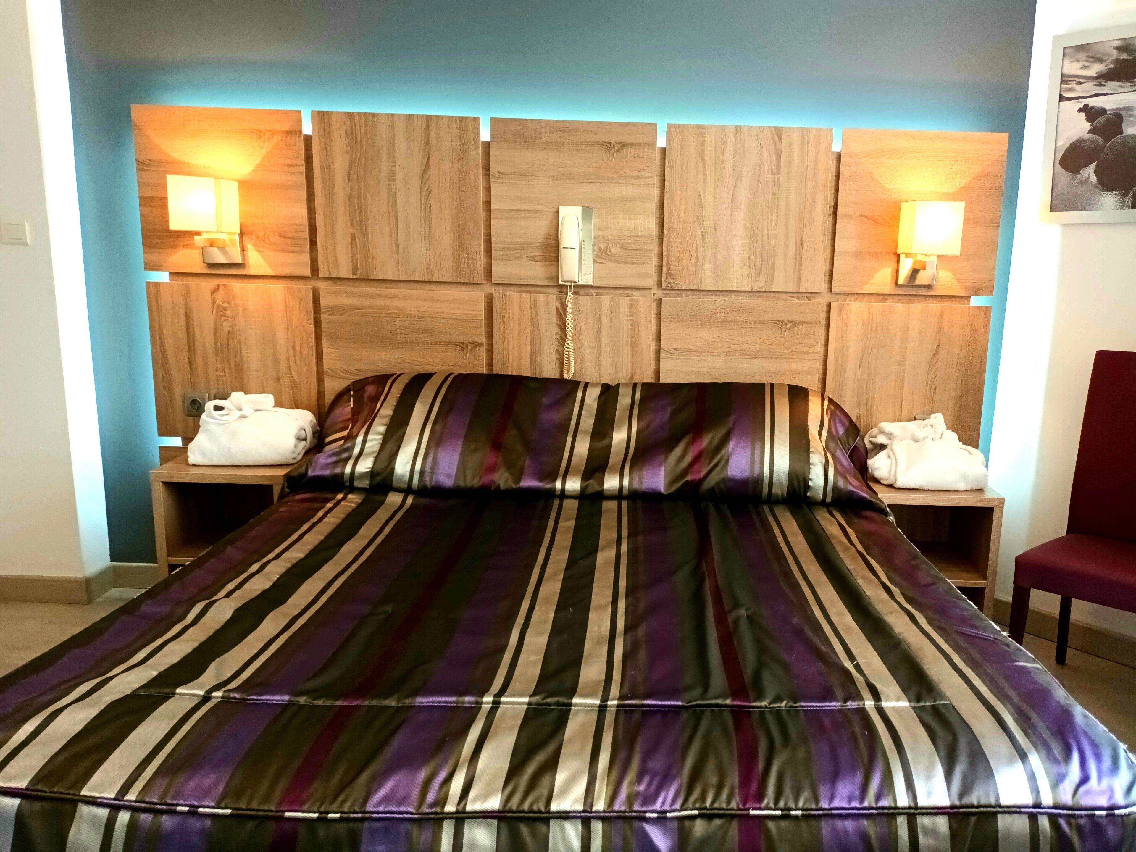 A hotel room with a wooden headboard, striped bedding, two bedside lamps, and a red chair.