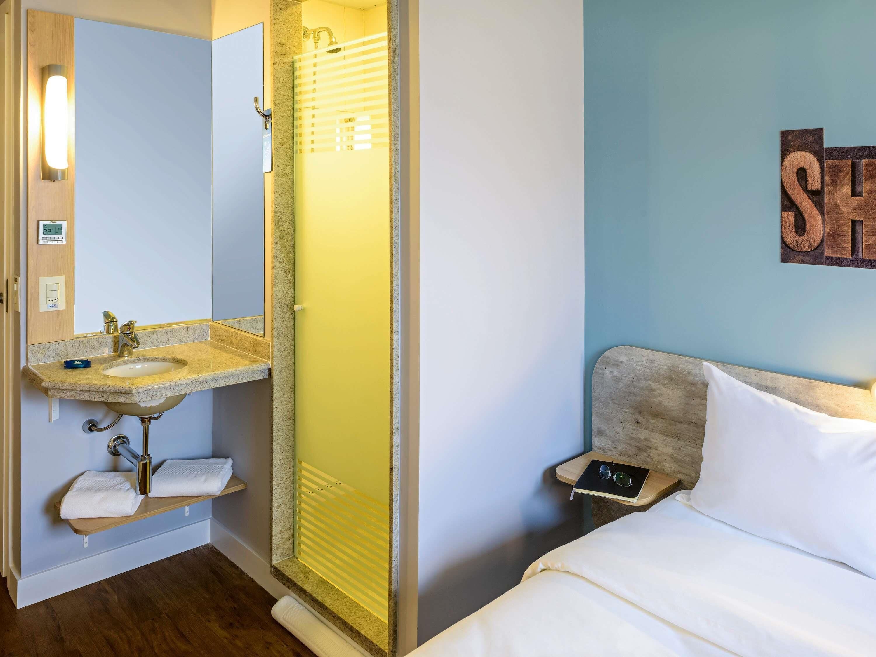 Habitación de hotel con baño que incluye lavamanos, espejo y ducha. Hay una cama con almohada y una mesita de noche con un libro y gafas.