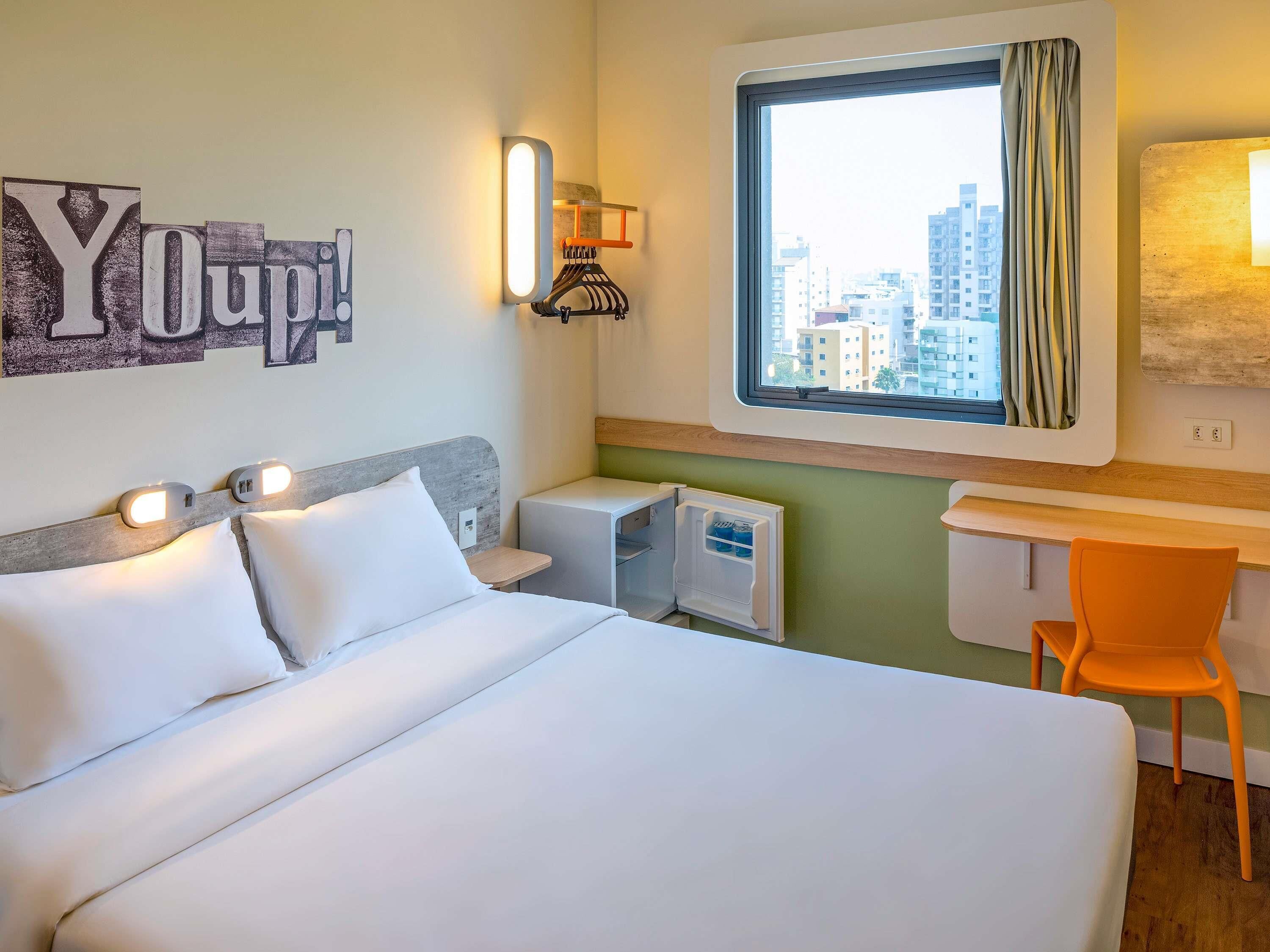 A hotel room with a bed, a desk, a chair, a window with a view of buildings, and a wall with illuminated letters.