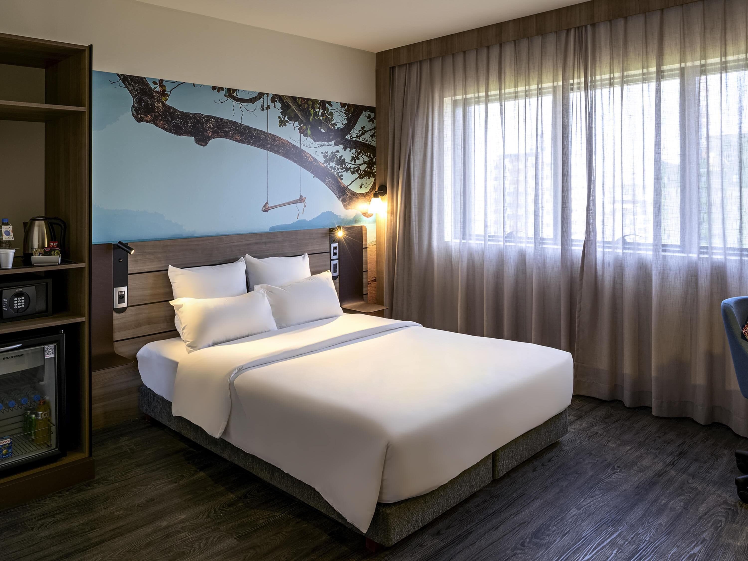 A hotel room with a bed, a nightstand, a wall-mounted TV, a shelf with a kettle, and a window with curtains.