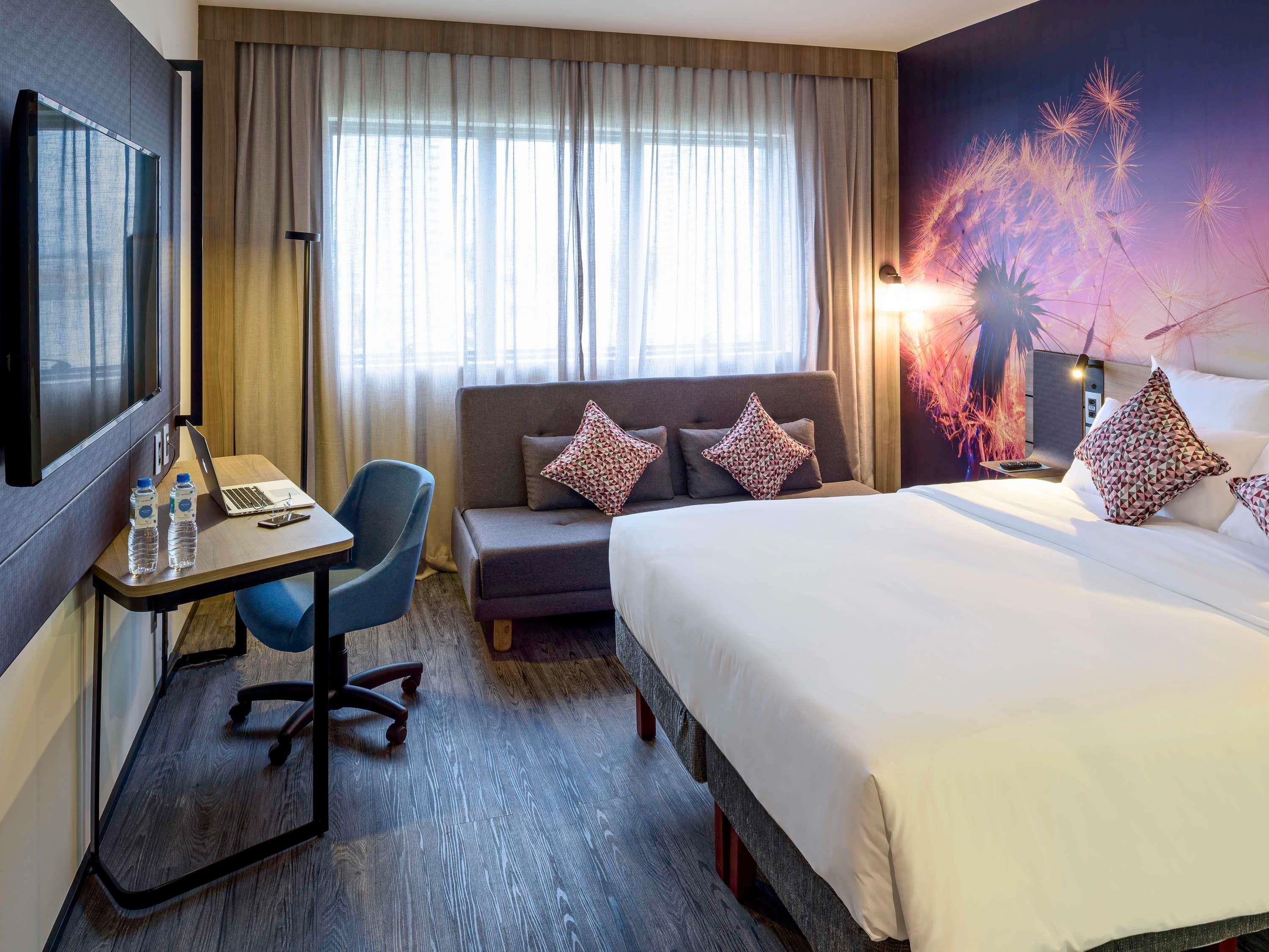 Habitación de hotel con cama, escritorio con una computadora portátil, silla, sofá y un ventanal con cortinas.